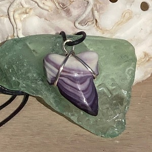 Puede incluir: Un colgante de concha de mar a rayas púrpuras y blancas en un collar de cordón negro. El colgante está envuelto en alambre plateado y descansa sobre un trozo de vidrio de mar verde.