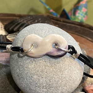 Könnte beinhalten: Ein schwarzes Lederarmband mit einem silbernen Unendlichkeits-Anhänger aus einer lila und weißen Muschel.