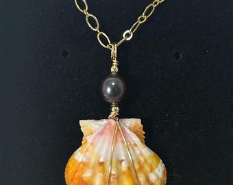 May include: A seashell pendant necklace with a gold chain. The shell is orange, yellow, and white, with gold wire wrapping. A dark gray pearl bead is above the shell. The necklace is displayed against a black background.