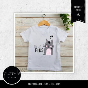 Op de afbeelding: Wit T-shirt met een cartoon kat ontwerp. De grijze kat heeft roze stippen op zijn buik en de woorden "Ich bin EINS" staan op het shirt. Het T-shirt hangt aan een houten hanger.