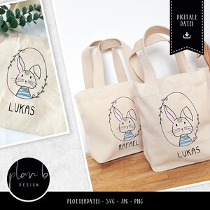 May include: Two white canvas tote bags with black bunny illustrations and the names "Lukas" and "Rafael" printed on them. The bags have black handles and are sitting on a wooden surface.