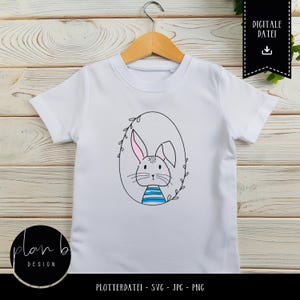 May include: White t-shirt featuring a black and white illustration of a bunny in an oval frame with leaves. The bunny has pink ears and blue and white stripes on its chest. The text "DIGITALE DATEI" is in a black box with a download icon.
