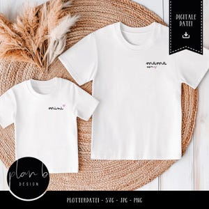 Könnte beinhalten: Zwei weiße T-Shirts mit dem Text "mama" und "mini" in schwarzer Schrift mit einem kleinen rosa Herz neben jedem Wort. Die Shirts liegen auf einer geflochtenen Strohmatte.