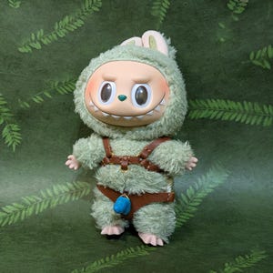 May include: A plush toy figure with a green furry costume and a smiling face. The figure has a brown leather harness and a blue charm. The background is a green surface with a fern pattern.