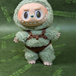 May include: A plush green monster doll with a smiling face, large eyes, and a furry hood. The doll has a brown harness and shorts. The background is a green surface with a fern pattern.