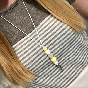 May include: A white lanyard with a yellow and gray silicone bead charm. The charm is shaped like a hexagon and has a silver clip.