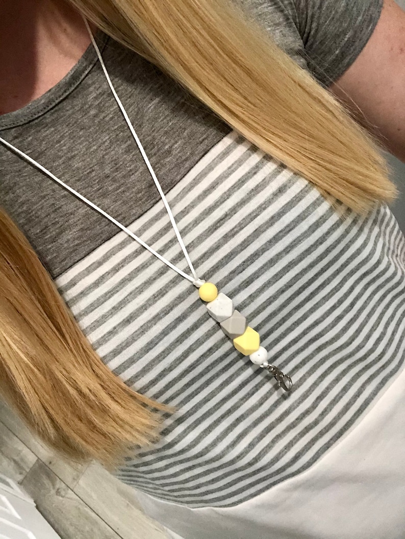 May include: A white lanyard with a yellow and gray silicone bead charm. The charm is shaped like a hexagon and has a silver clip.