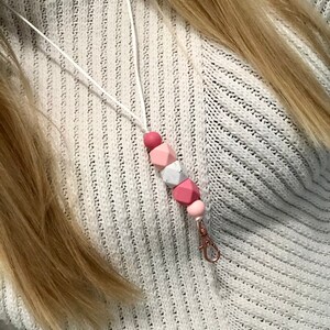 May include: A white lanyard with a pink and white silicone bead charm. The charm has a geometric shape and is attached to a metal clip.