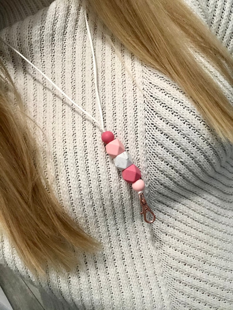 May include: A white lanyard with a pink and white silicone bead charm. The charm has a geometric shape and is attached to a metal clip.