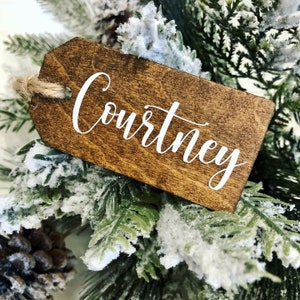 May include: A wooden Christmas ornament with the name "Courtney" written in white cursive lettering. The ornament is brown and has a rustic look. It is hanging on a snowy evergreen branch.