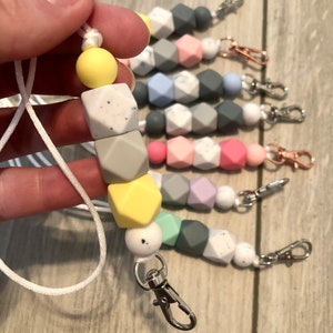 May include: A white cord with a silver clip and a string of colorful silicone beads. The beads are in various shades of gray, yellow, green, and pink. The beads have a speckled pattern.