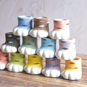 May include: A set of 12 ceramic cat paw planters in various colours, including blue, green, yellow, pink, and brown. The planters are arranged in a pyramid shape and have a white base.