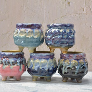 Puede incluir: Cinco pequeñas macetas de cerámica con un acabado de esmalte de goteo. Las macetas son en tonos de azul, morado, rosa y negro. Cada maceta tiene tres pequeños pies.