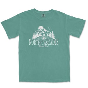 May include: A teal blue t-shirt with a white graphic of mountains, a lake, and trees. The text "NORTH CASCADES National Park" is printed below the graphic.