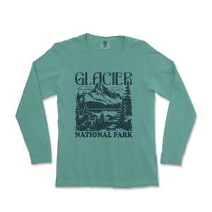 Könnte beinhalten: Ein türkisfarbenes Langarmshirt mit einer Grafik im Vintage-Stil einer Bergkette und dem Text "GLACIER NATIONAL PARK".