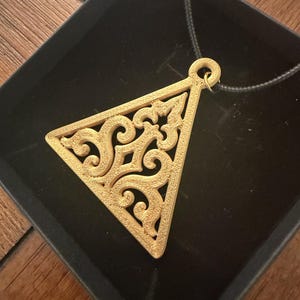 May include: A gold-colored triangular pendant featuring an intricate, symmetrical filigree cutout pattern. The pendant has a textured surface and is attached to a thin black cord, resting inside a black square box with a velvet-like interior lining.