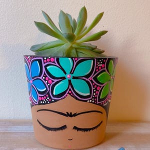 May include: A hand-painted terracotta flower pot featuring a face design with closed eyes and a floral crown. The pot is filled with a green succulent plant. The floral crown has blue, teal, pink, and green flowers.