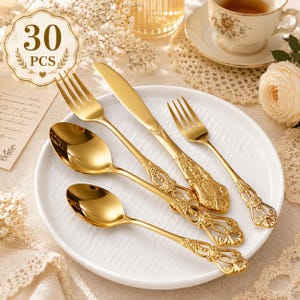 May include: A set of gold-colored stainless steel flatware featuring ornate, embossed handles with intricate floral detailing. The collection includes a dinner fork, a dinner knife, a smaller fork, and two spoons of varying sizes, all arranged on a white plate.