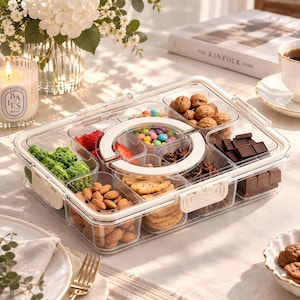 May include: A rectangular clear plastic food storage container with multiple compartments and a white circular center insert. The container is filled with various snacks including broccoli, strawberries, almonds, cookies, walnuts, chocolate, and candies.