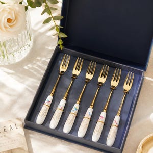 May include: A set of six gold-toned metal dessert forks with white ceramic handles. Each handle features a delicate floral pattern with small colorful accents and a gold-speckled design. The forks are arranged neatly inside a dark blue presentation box.