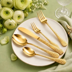May include: A set of gold-toned flatware including a dinner fork, a smaller fork, a dinner knife, a large spoon, and a small spoon, all featuring an intricate embossed scroll pattern on the handles, arranged on a white plate.