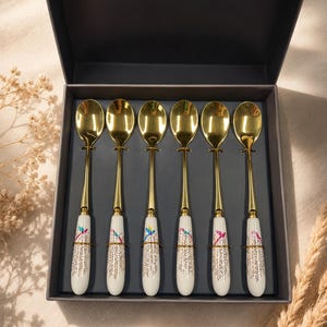 May include: A set of six gold-toned metal spoons arranged in a dark gray presentation box. Each spoon features a white ceramic handle decorated with a colorful hummingbird illustration and a gold-colored floral pattern at the base of the handle.