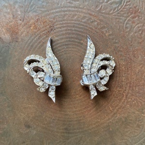 May include: A pair of silver tone earrings with a floral design. The earrings are adorned with clear rhinestones and have a delicate, feminine look.