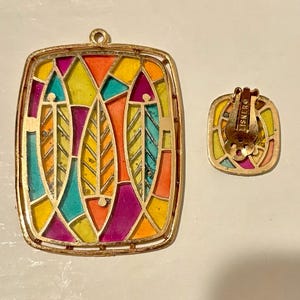 Könnte beinhalten: Ein goldfarbenes Anhänger- und passendes Clip-Ohrring-Set. Der Anhänger und der Ohrring weisen ein farbenfrohes Buntglasdesign mit gelben, orangefarbenen, türkisfarbenen und violetten Farbtönen auf. Der Ohrring ist mit "LISNER" gekennzeichnet.