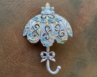 May include: A silver-toned brooch in the shape of an umbrella with a bow at the bottom. The umbrella is decorated with clear and blue rhinestones.