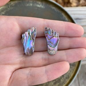May include: Two silver-toned foot-shaped earrings with iridescent abalone shell inlays. The earrings display a range of colors, including blue, purple, and green, and are set against a neutral background.