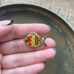 May include: A gold-colored metal pin shaped like a fish with the word "JESUS" written inside. The letters are in different colors: red, yellow, green, and black.