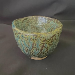 Op de afbeelding: Een kleine, groene en bruine keramische kom met een getextureerd, organisch patroon. De kom is licht onregelmatig van vorm en heeft een afgeronde bodem.