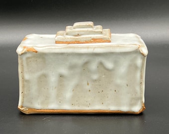 Puede incluir: Una caja rectangular de cerámica con una tapa que presenta un asa escalonada. La pieza tiene un acabado de esmalte grueso de color blanco roto que gotea por los lados, revelando una arcilla de tono marrón rojizo en los bordes y la base.