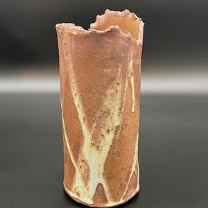 Puede incluir: Un jarrón cilíndrico de cerámica con un borde superior rugoso e irregular. El exterior presenta una superficie texturizada de color marrón tierra con vetas irregulares de esmalte crema que recorren verticalmente el cuerpo del objeto.