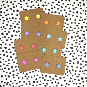 May include: Assortment of colourful flower-shaped stud earrings displayed on brown card stock. The earrings are in shades of yellow, orange, pink, purple, blue, and turquoise.