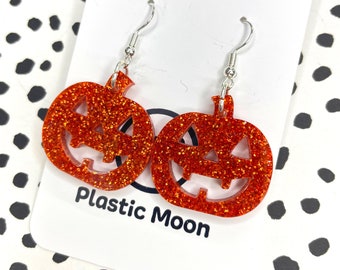 May include: A pair of orange glitter pumpkin earrings with a jack-o'-lantern face. The earrings are on a white card with the text "Plastic Moon".