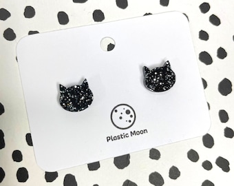 以下が含まれることがあります： 白いカードに「Plastic Moon」の文字が書かれた、黒いグリッターの猫の顔のスタッドピアスのペアです。