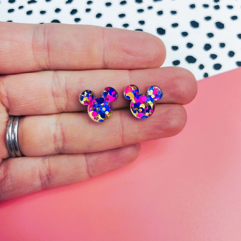Puede incluir: Un par de pendientes de bot&oacute;n dorados con un dise&ntilde;o de confeti en rosa, azul y dorado. Los pendientes tienen forma de orejas de Mickey Mouse.