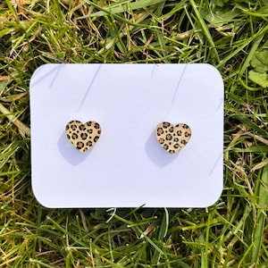 May include: A pair of heart-shaped stud earrings featuring a leopard print design. The earrings are presented on a white card, set against a backdrop of green grass. The earrings are made of wood.