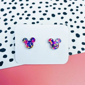 Puede incluir: Un par de pendientes de bot&oacute;n con una silueta de Mickey Mouse llena de confeti de colores. El confeti es rosa, azul, amarillo y dorado.