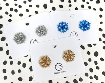 May include: Three pairs of snowflake stud earrings on white cards. The earrings are silver, gold, and blue with glitter. The cards have the text "Plastic Moon" printed on them.