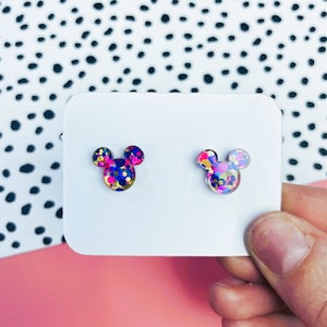 Puede incluir: Un par de pendientes de bot&oacute;n con forma de cabeza de Mickey Mouse. Los pendientes est&aacute;n llenos de confeti de colores y tienen un contorno dorado.