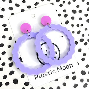 May include: A pair of lavender hoop earrings with a scalloped edge and a pink accent on the top. The earrings are on a white card with the text "Plastic Moon".