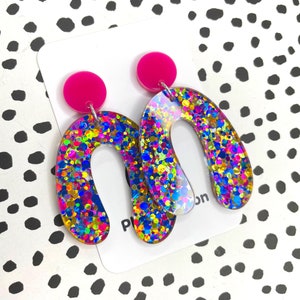 Peut inclure: Une paire de boucles d'oreilles créoles en paillettes roses et arc-en-ciel. Les boucles d'oreilles sont en forme de fer à cheval et ont un dos rose.