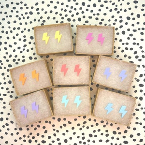 May include: Eight small boxes with different colored lightning bolt earrings. The boxes are arranged in two rows of four. The earrings are in the colors yellow, pink, red, blue, light blue, purple, orange, and light orange.
