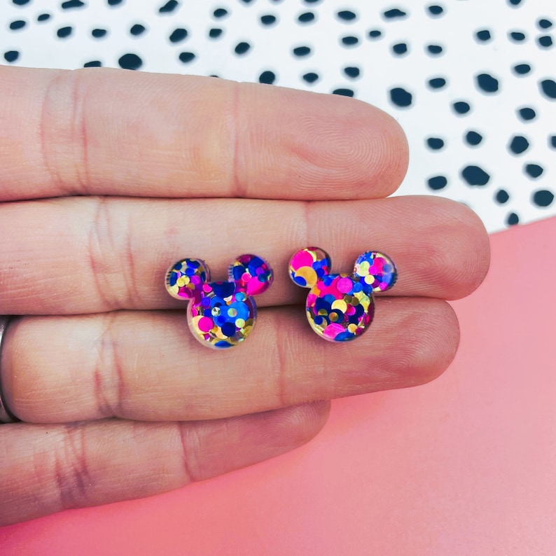 Puede incluir: Un par de pendientes de resina transparente con forma de Mickey Mouse con confeti de colores en su interior. El confeti es rosa, azul, dorado y amarillo.