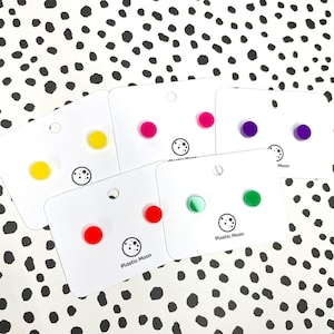 May include: Six pairs of colourful stud earrings displayed on white cards. The cards have the text "Plastic Moon" printed on them. The earrings are in the colours red, yellow, green, pink, purple, and a clear green with a white swirl.