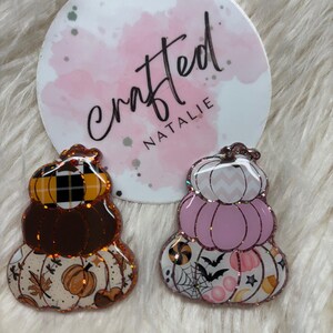 May include: Two stacked pumpkin-shaped pins with glitter edges. One features a plaid pattern, brown, and autumn leaves. The other has a pink and white chevron pattern, bats, and spiderwebs. A round logo with the text "crafted NATALIE" is in the background.