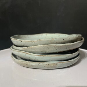 May include: A stack of four round ceramic plates with an irregular, hand-formed edge. The plates feature a light mint green glaze with small dark brown speckles throughout the surface, showing a smooth, glossy finish and a natural, organic shape.
