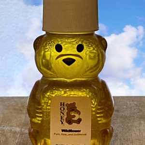 May include: A clear glass bear-shaped bottle filled with honey. The bottle has a white label with the text "HONEY Wildflower Pure, Raw, and Unfiltered Net Wt. 2 oz (56.70 gm)".